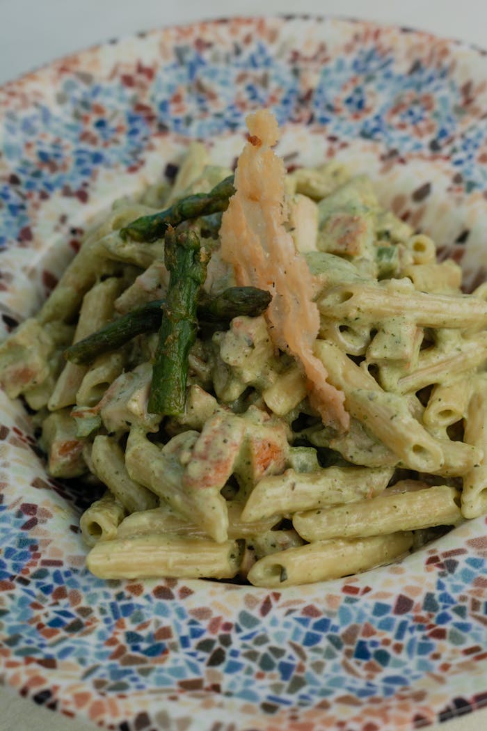 portfolio-05 Savor this creamy pesto penne pasta garnished with asparagus and a parmesan crisp on a mosaic plate.